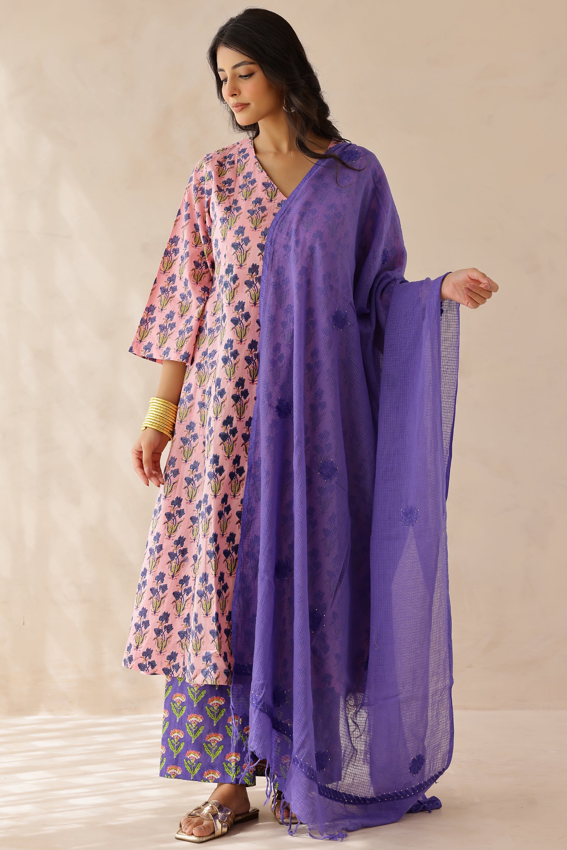 Woman wearing a pink printed cotton kurta with a V-neckline and 3/4th sleeves, paired with purple printed palazzo pants and a contrasting kota doria dupatta