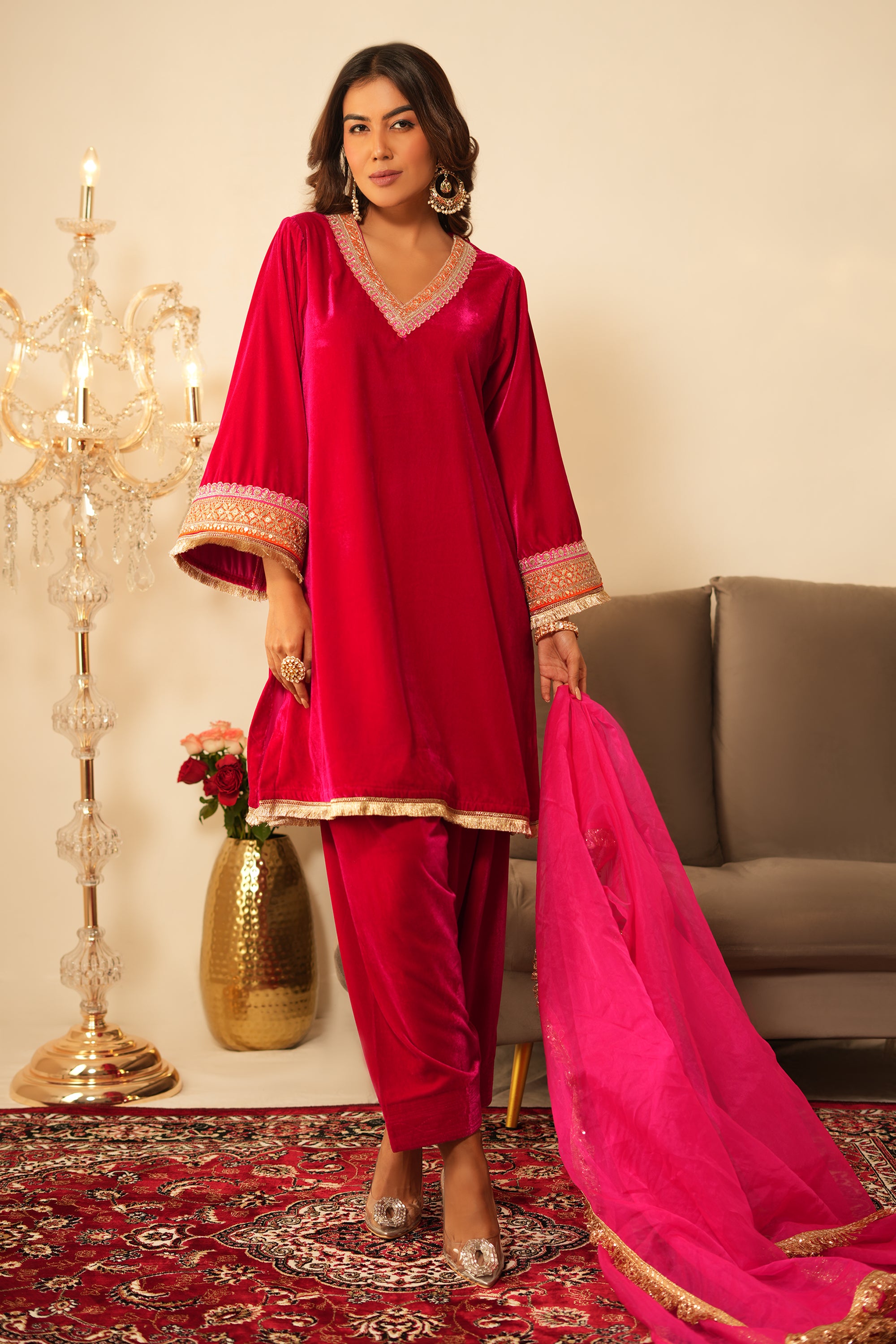 Woman in a traditional pink velvet outfit with pink dupatta standing in a decorated room.