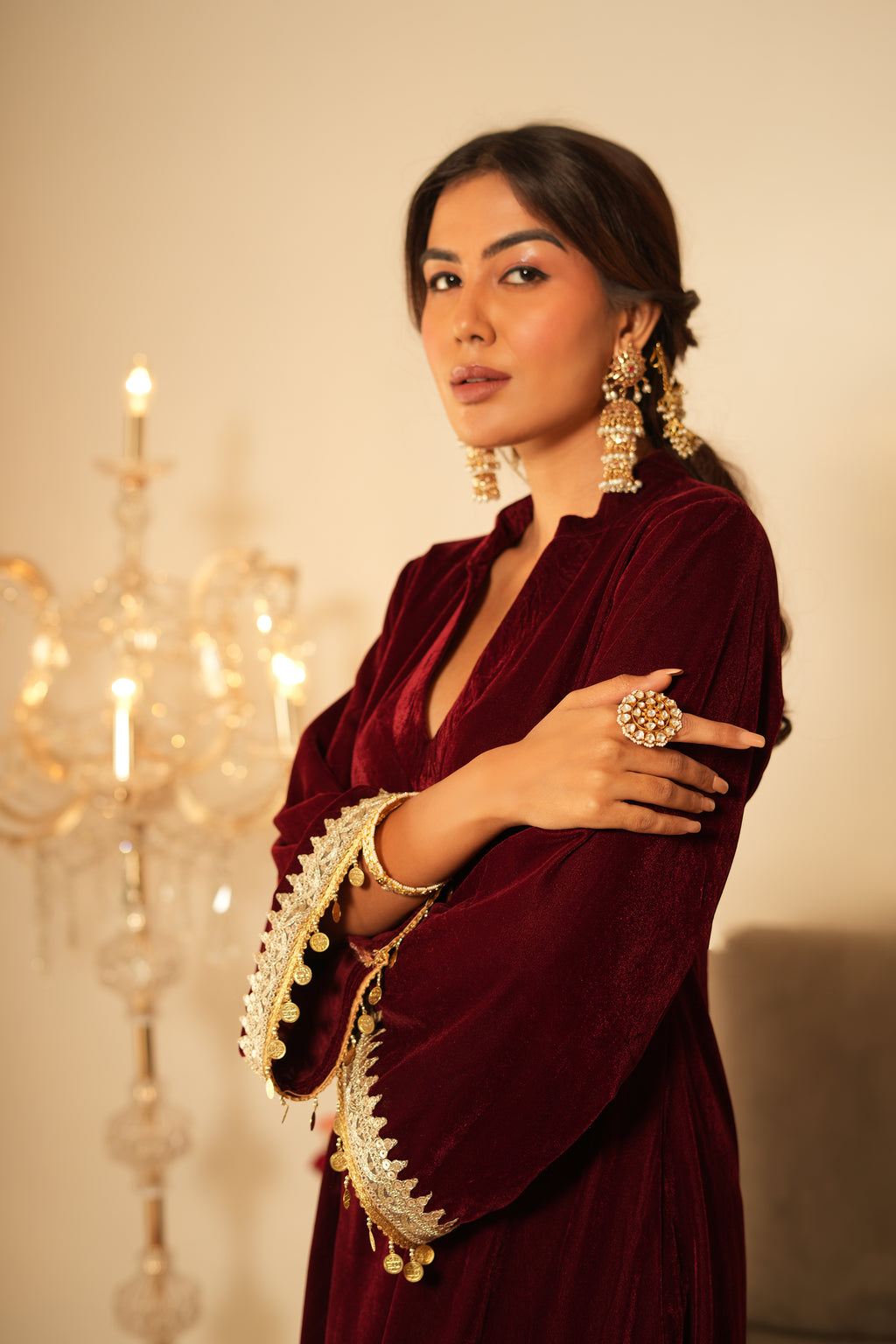 Woman in a maroon traditional outfit with gold details, standing in front of a chandelier.