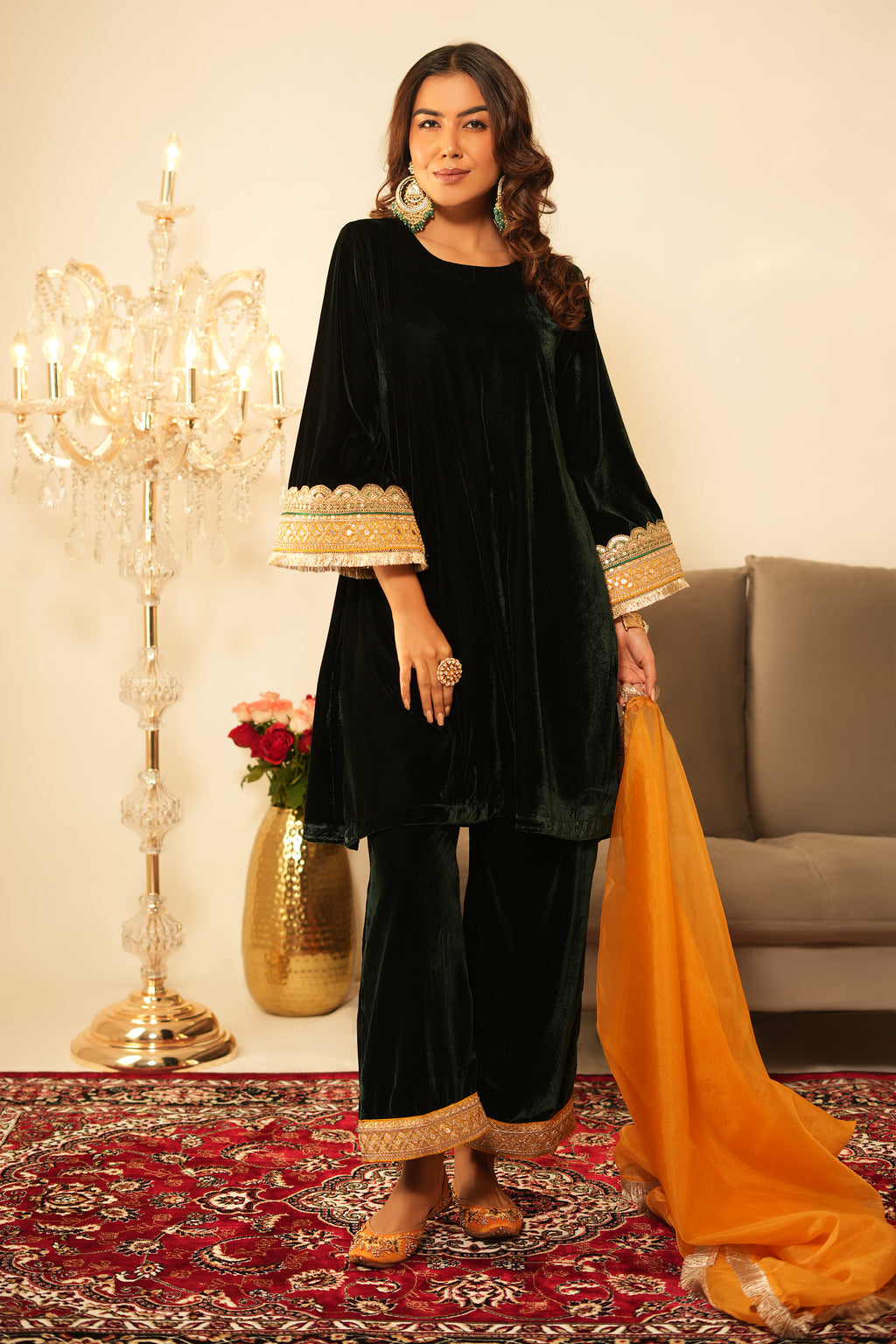 Woman in black traditional outfit with gold trim standing in a decorated room with a chandelier and sofa.