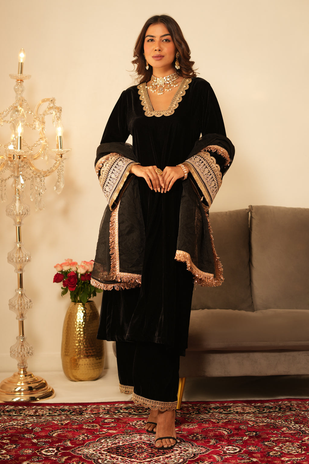 Woman in a black traditional outfit with gold embroidery standing in a room with a chandelier and flowers.