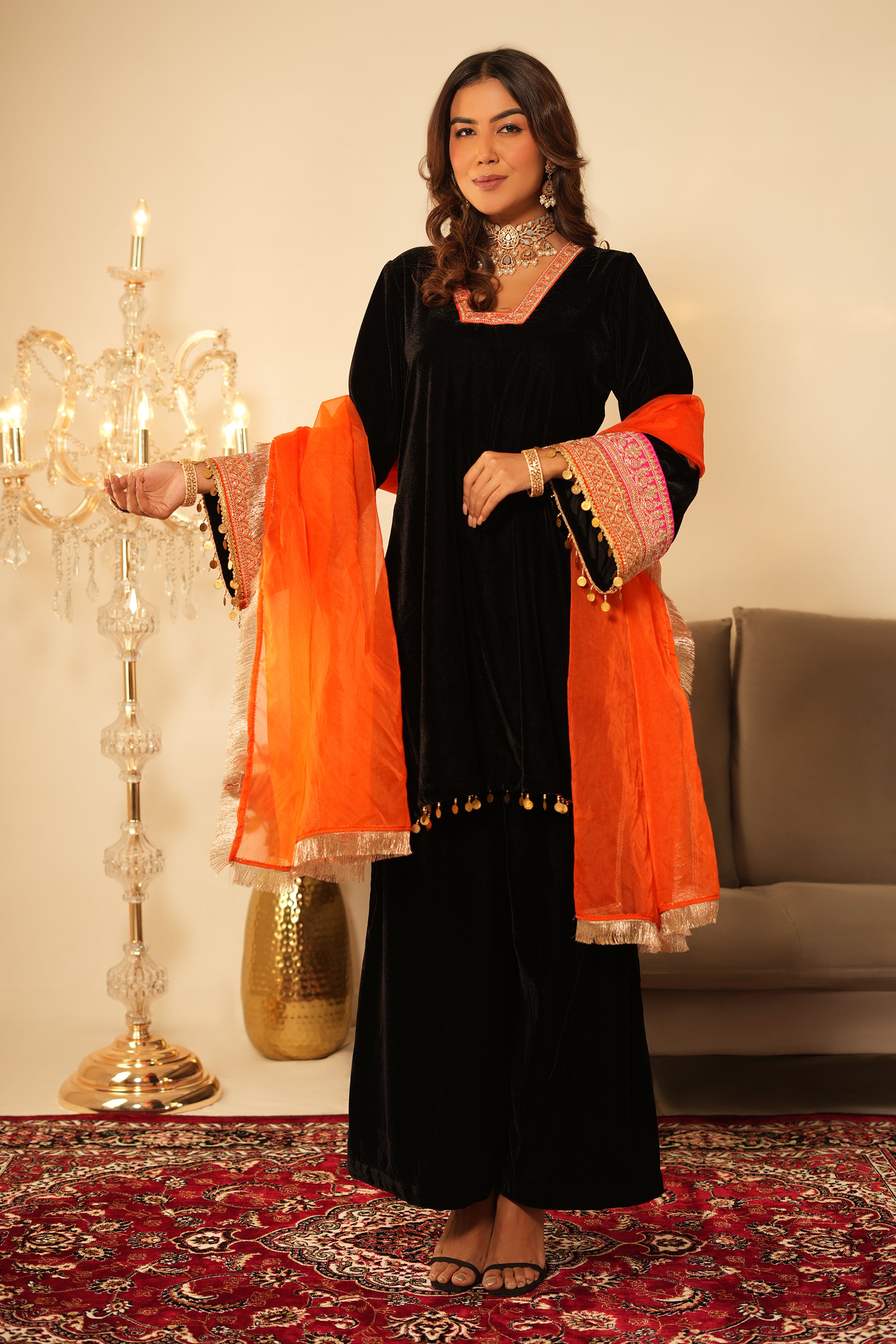 Woman in black traditional outfit with orange dupatta standing in a decorated room.