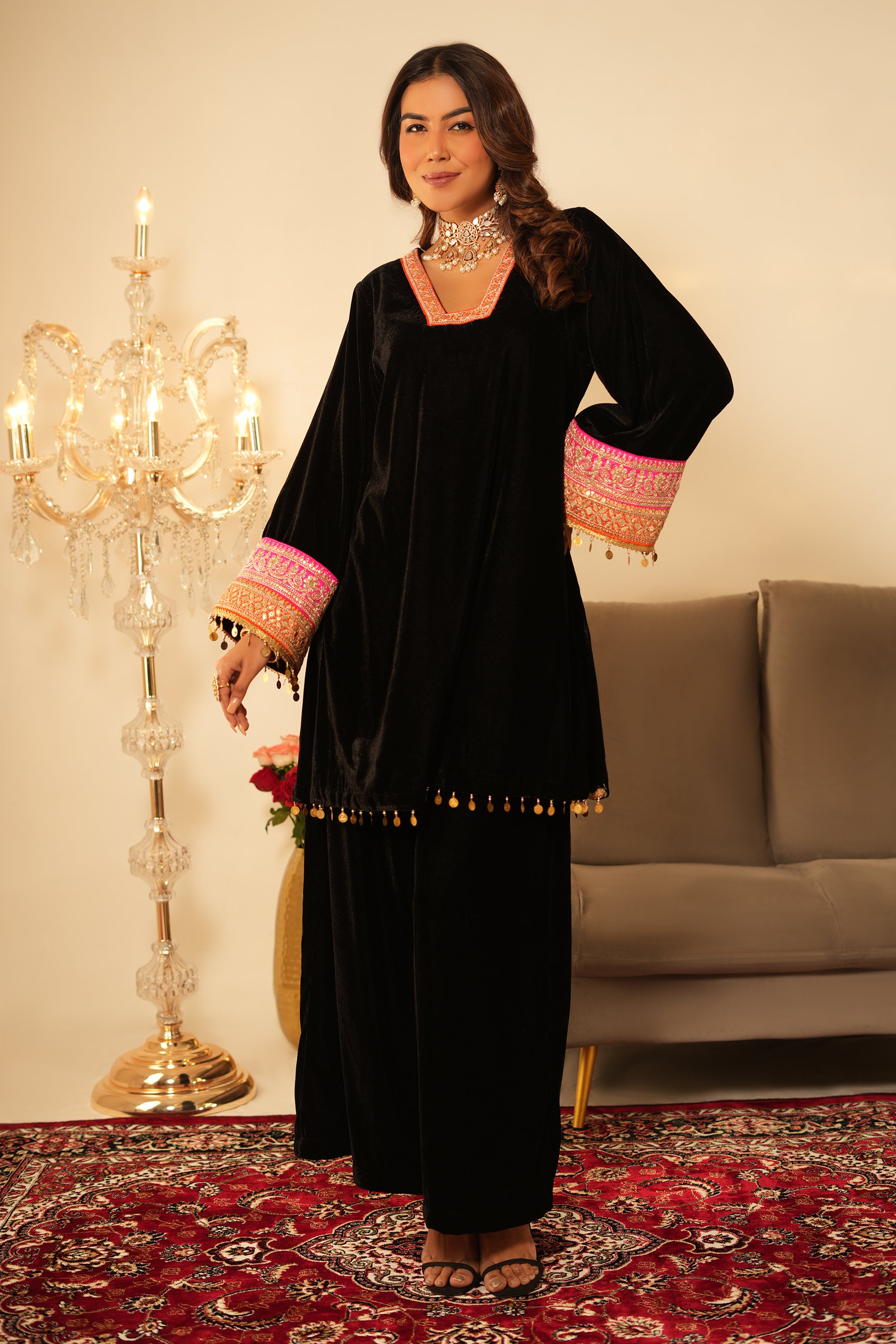 Woman in a black traditional outfit with pink and gold details standing on a red patterned rug.