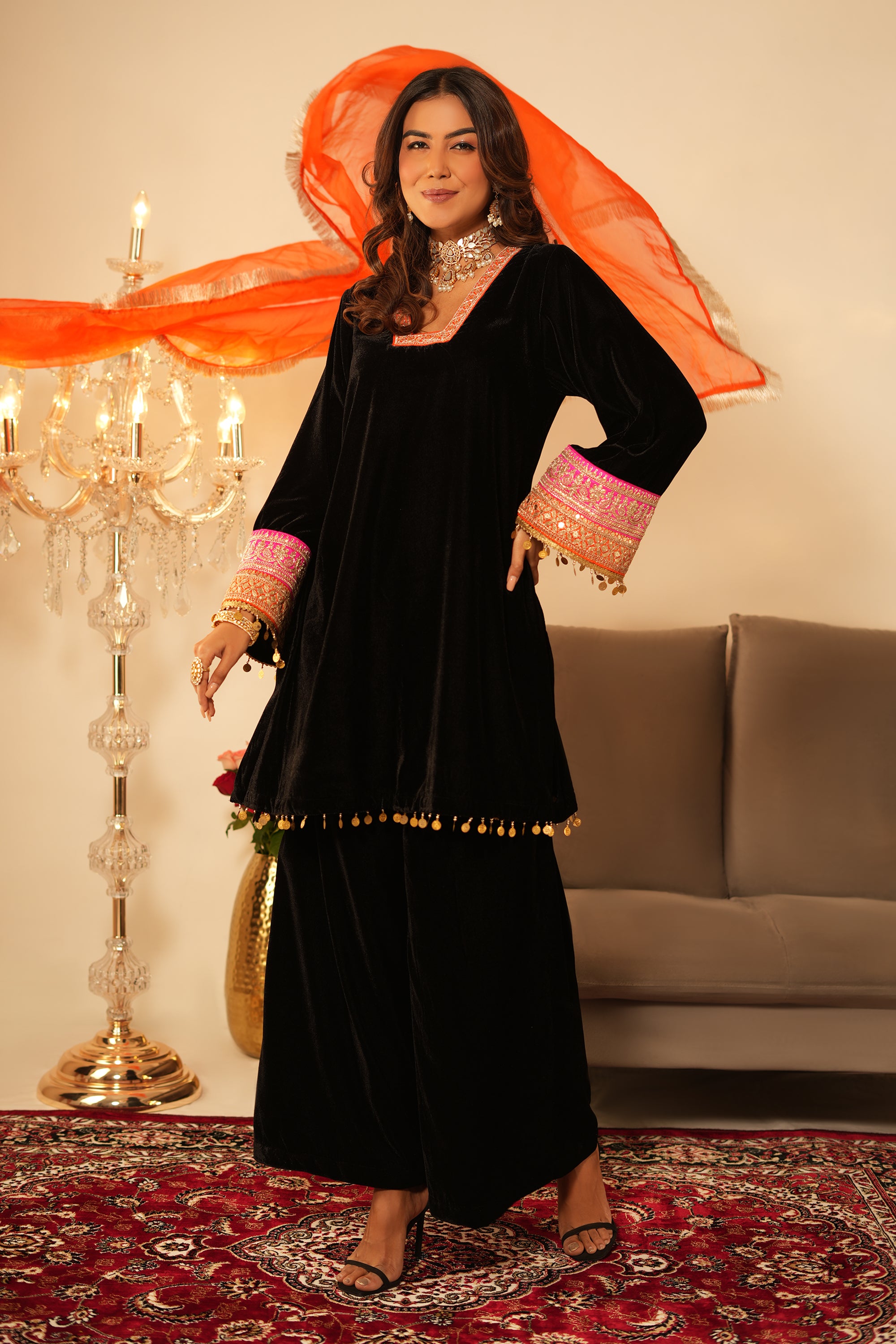 Woman in black traditional outfit with orange dupatta in a decorated room.