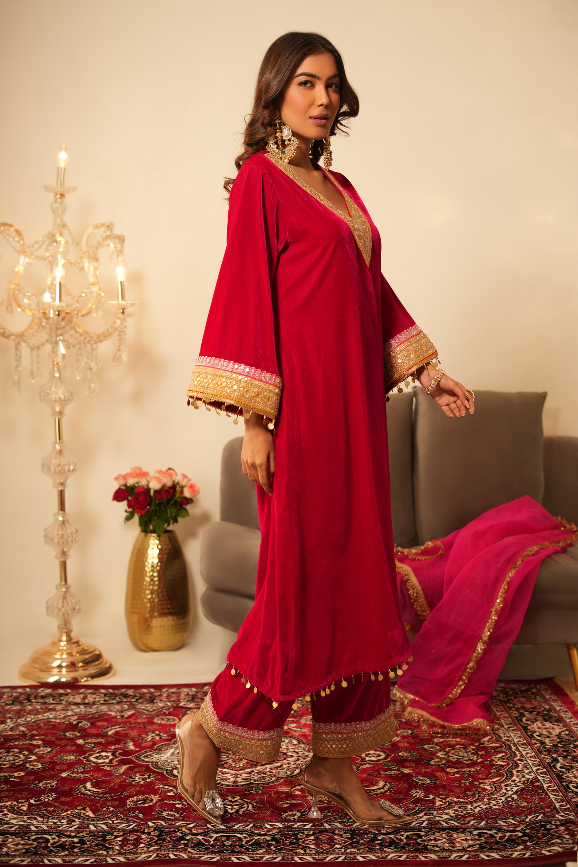 Woman in a pink traditional velvet kurta with gold details standing in a decorated room.