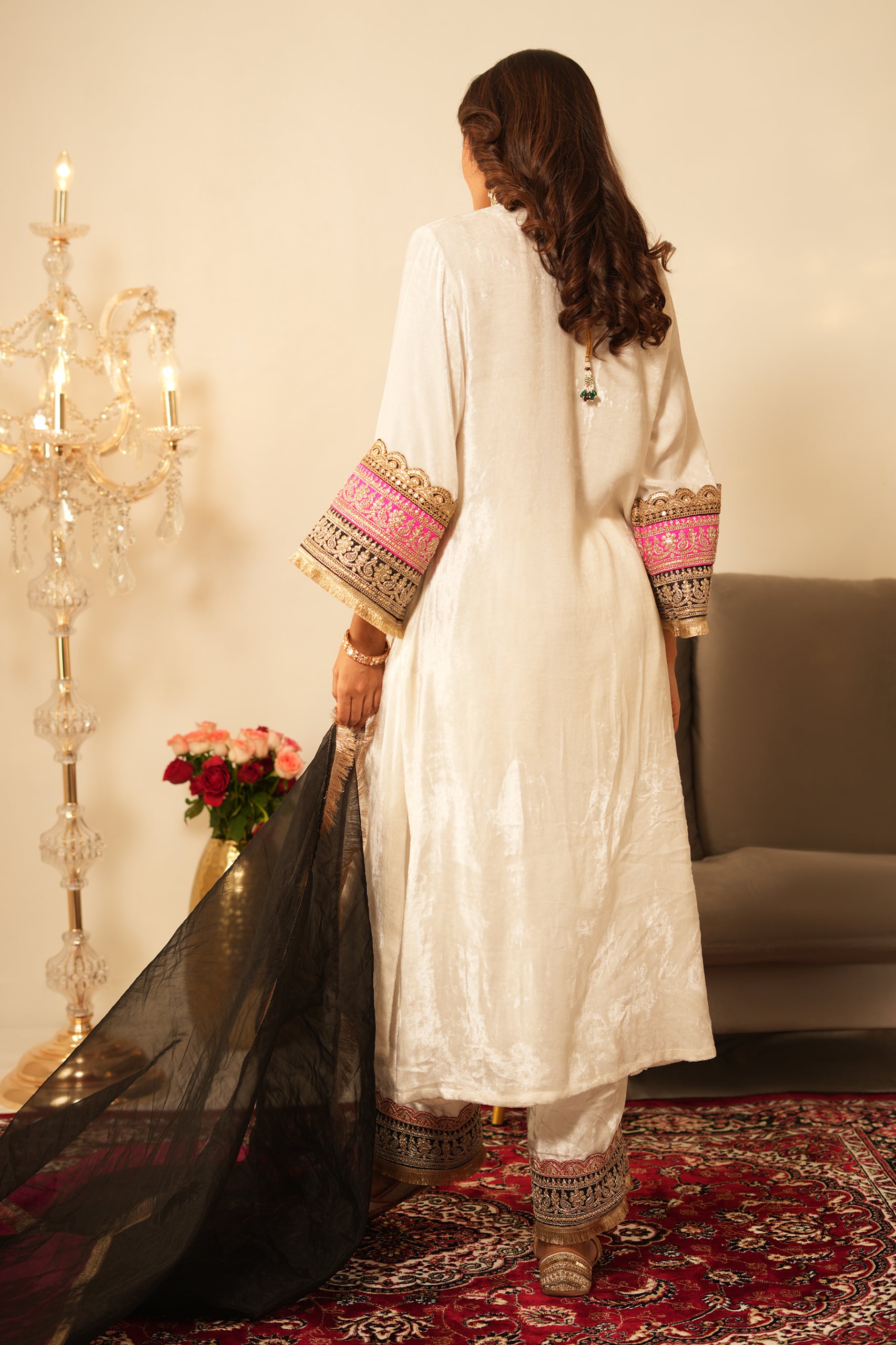 Woman in a white traditional velvet kurta with gold details standing in a decorated room with black and pink organza dupatta