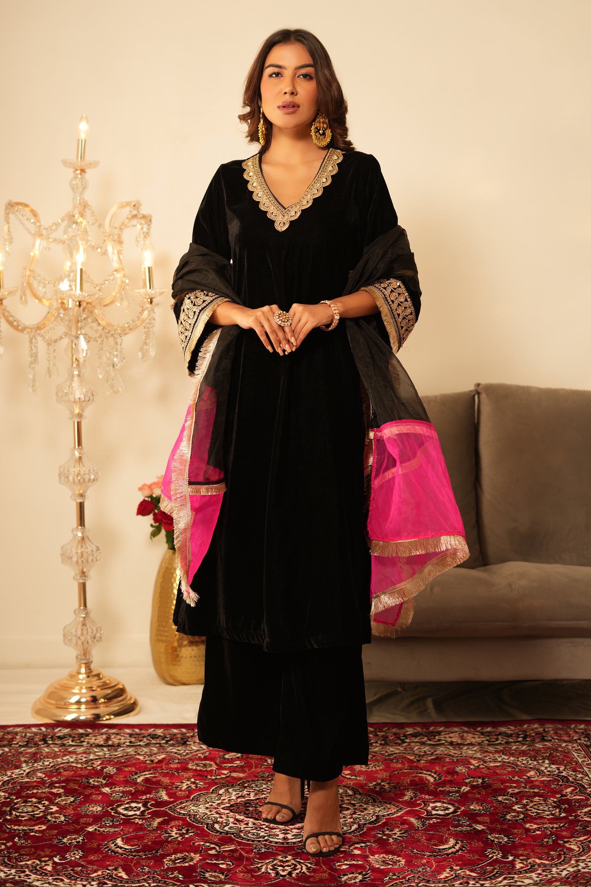 Woman in a black velvet kurta outfit with gold trim, wearing a black and pink organza dupatta standing in a room with  decorative items.