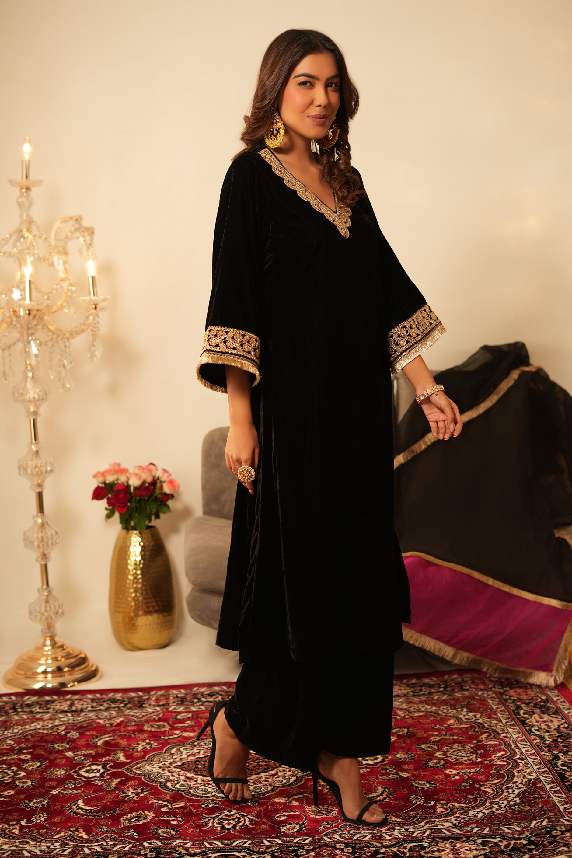 Woman in a black velvet kurta outfit with gold trim, wearing a black and pink organza dupatta standing in a room with  decorative items.