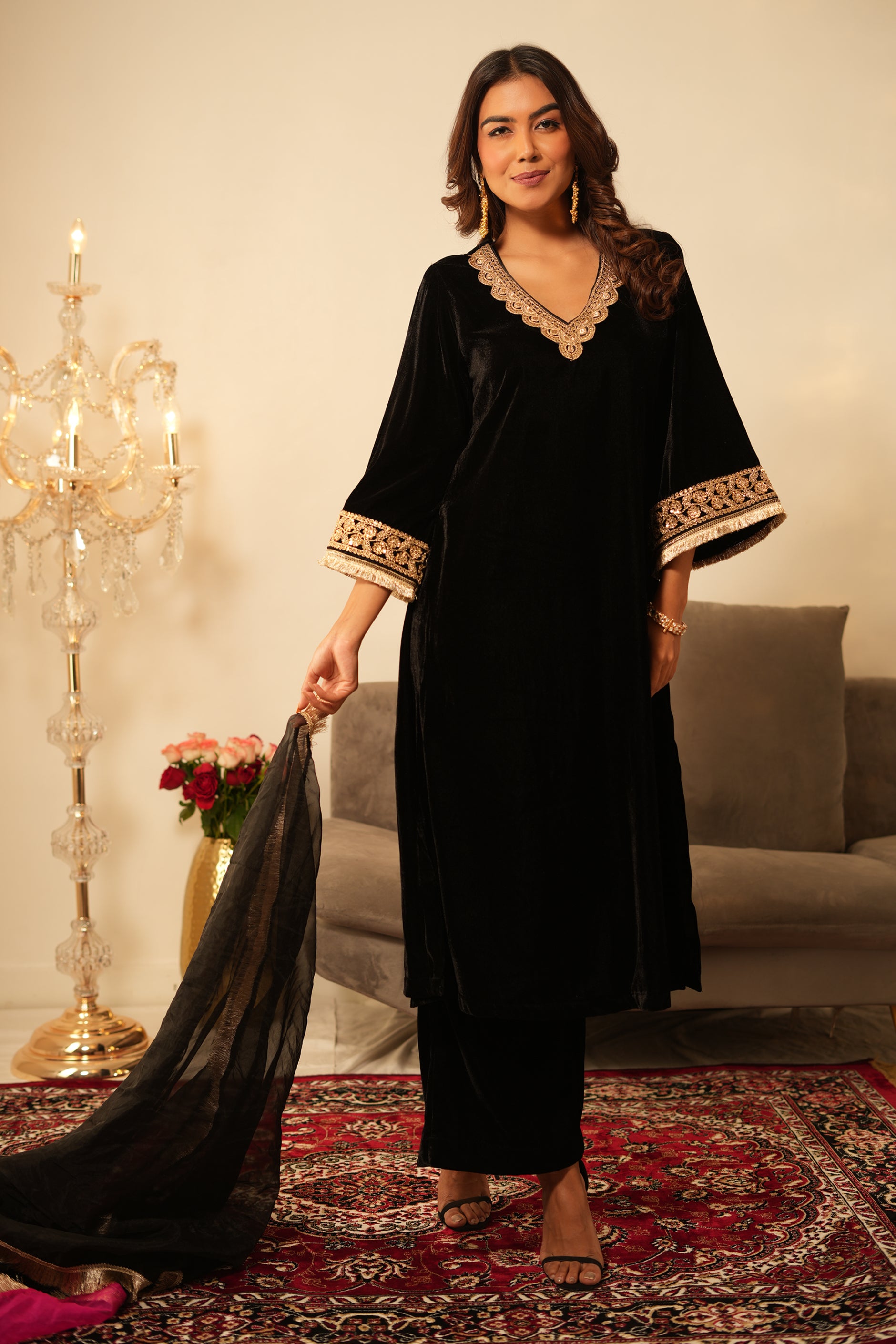 Woman in a black velvet kurta outfit with gold trim, wearing a black and pink organza dupatta standing in a room with  decorative items.
